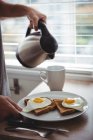 Homem segurando sua placa de café da manhã enquanto derramando água quente em caneca na cozinha — Fotografia de Stock