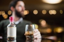 Крупный план маленькой бутылочки ликера на столе в баре с человеком, держащим стакан напитка на заднем плане — стоковое фото