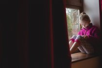 Fille assise sur le rebord de la fenêtre et en utilisant une tablette numérique à la maison — Photo de stock