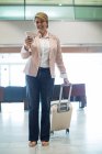 Femme d'affaires souriante avec sac trolley utilisant un téléphone portable dans la salle d'attente au terminal de l'aéroport — Photo de stock