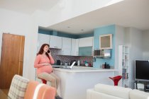 Mujer embarazada hablando por teléfono móvil en la cocina en casa - foto de stock