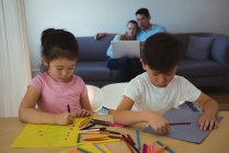 Хлопчик і дівчинка малюють в папері, а батьки використовують ноутбук у фоновому режимі вдома — стокове фото
