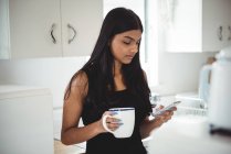 Frau benutzt Handy, während sie zu Hause in der Küche Kaffee trinkt — Stockfoto