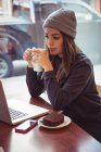 Woman in winter clothing holding coffee cup and looking at laptop in restaurant — Stock Photo