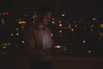 Человек, использующий свой мобильный телефон возле окна жалюзи ночью — стоковое фото