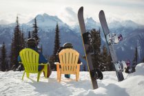 Vista trasera de la pareja sentada en silla por tablas de snowboard en la montaña cubierta de nieve - foto de stock