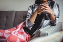 Seção média de mulher segurando xícara de café sentado no sofá na sala de estar em casa — Fotografia de Stock