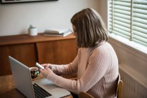 Femme utilisant un téléphone mobile tout en travaillant sur un ordinateur portable à la maison — Photo de stock