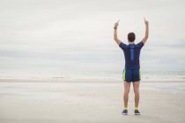 Rear view of athlete standing on beach with hands raised — Stock Photo