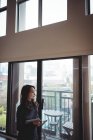 Frau hält digitales Tablet in der Hand, während sie zu Hause durchs Fenster schaut — Stockfoto