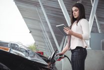 Schöne Frau mit digitalem Tablet beim Laden von Elektroautos an der Ladestation — Stockfoto