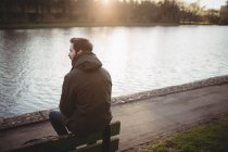 Nachdenklicher junger Mann sitzt auf Bank am Flussufer — Stockfoto