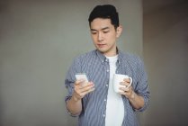 Hombre usando el teléfono móvil mientras toma una taza de café en la oficina - foto de stock
