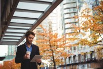 Бизнесмен с одноразовой чашкой кофе и с помощью цифрового планшета во время прогулки по улице — стоковое фото
