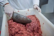 Sección media del carnicero utilizando cuchara para eliminar la carne picada del contenedor en la fábrica de carne - foto de stock