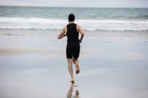 Вид сзади на человека в купальнике и плавательной шапочке, бегущего по пляжу — стоковое фото