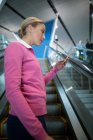 Comutador feminino usando telefone celular na escada rolante no aeroporto — Fotografia de Stock
