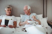 Hombre mayor desayunando mientras mujer leyendo un libro en la cama en la habitación - foto de stock