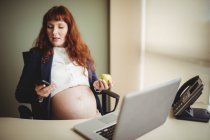 Беременная деловая женщина, использующая мобильный телефон, имея яблоко в офисе — стоковое фото