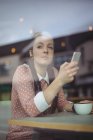 Задумчивая женщина, пользующаяся мобильным телефоном за чашечкой кофе в кафе — стоковое фото