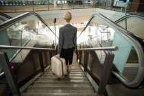 Вид сзади на деловую женщину, стоящую на эскалаторе с багажом в аэропорту — стоковое фото