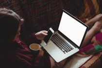 Женщина пьет кофе с помощью ноутбука и мобильного телефона на диване дома — стоковое фото