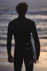 Vue arrière d'un homme portant une planche de surf debout sur la plage au crépuscule — Photo de stock