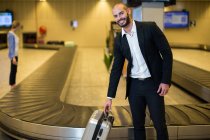 Портрет улыбающегося бизнесмена с тележкой в зоне ожидания терминала аэропорта — стоковое фото