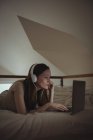 Woman with headphones using laptop on bed at home — Stock Photo