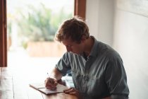 Мужчина сидит за столом и пишет на блокноте дома — стоковое фото
