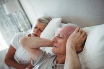 Старший чоловік лежить на ліжку і прикриває вуха руками в спальні — стокове фото