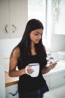 Mujer usando el teléfono móvil mientras sostiene la taza de café en la cocina en casa - foto de stock
