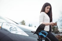 Belle femme utilisant un téléphone mobile tout en chargeant une voiture électrique sur la voiture de rue — Photo de stock