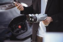 Mid section of man using mobile phone while holding car charger at electric vehicle charging station — Stock Photo
