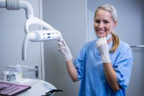 Assistant dentaire souriant à la caméra à côté de la lumière à la clinique dentaire — Photo de stock