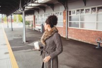 Mulher segurando copo de café descartável enquanto em pé na plataforma estação ferroviária — Fotografia de Stock