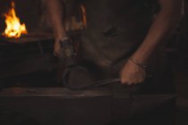 Section médiane du forgeron tenant une fer à cheval en métal avec des pinces sur l'enclume à l'aide d'un marteau pour former — Photo de stock