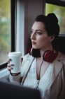 Mujer reflexiva sosteniendo taza de café desechable por ventana en tren - foto de stock