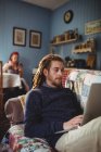 Hipster homme utilisant un ordinateur portable tout en étant assis sur le canapé avec une femme en arrière-plan à la maison — Photo de stock