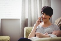 Mother talking by smartphone while baby sleeping in her arm at living room — Stock Photo