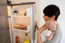 Mãe com seu bebê olhando para a geladeira na cozinha em casa — Fotografia de Stock