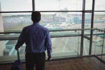 Вид сзади бизнесмена с багажом, смотрящего через стеклянное окно в аэропорту — стоковое фото