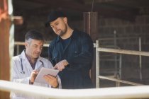 Працівник ферми вказує на планшетний комп'ютер під час обговорення з ветеринаром у сараї — стокове фото