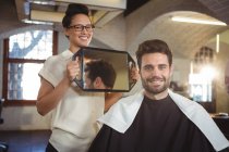 Sorridente parrucchiere femminile che mostra all'uomo il suo taglio di capelli nello specchio al salone — Foto stock