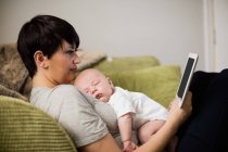 Mutter nutzt digitales Tablet, während Baby im heimischen Wohnzimmer auf ihr schläft — Stockfoto