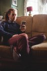 Jovem usando telefone celular enquanto se senta no sofá em casa — Fotografia de Stock