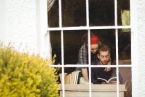 Pareja leyendo novela vista a través de ventana en casa - foto de stock