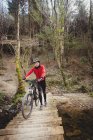 Vorderansicht des Mountainbikers, der mit dem Fahrrad auf einer Fußgängerbrücke im Wald wandert — Stockfoto