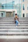 Full length of young woman holding phone while sitting on steps — Stock Photo