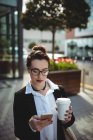 Молодая деловая женщина с одноразовой чашкой кофе с помощью мобильного телефона на улице — стоковое фото